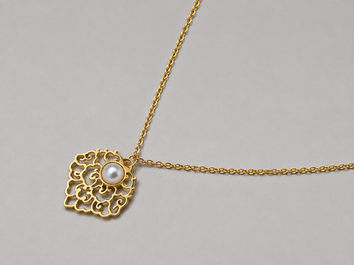A gold filigree pendant necklace featuring a central white pearl, displayed against a neutral gray background.